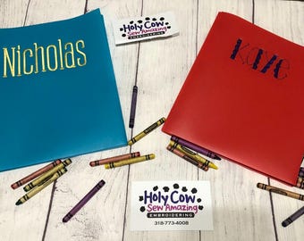 May include: Two personalized notebooks, one teal with "Nicholas" in gold lettering and one red with "Kaye" in blue lettering. Crayons are scattered around the notebooks. A business card for "Holy Cow Sew Amazing Embroidery" is also present.