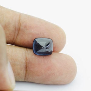 May include: A dark blue gemstone with a faceted cut, held between two fingers.