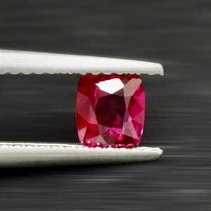 May include: A cushion-cut, faceted ruby gemstone held by tweezers against a dark background. The ruby is a vibrant red color with multiple facets reflecting light. The tweezers are silver and have a serrated edge.