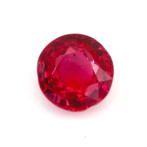 Puede incluir: Una gema de rubí tallada en forma redonda y facetada. La gema es de un color rojo intenso y vibrante, con facetas internas visibles que capturan y reflejan la luz. El rubí está sobre un fondo blanco liso.