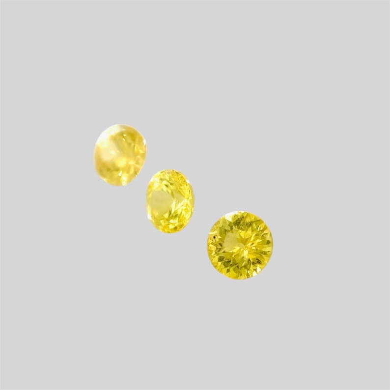 May include: Three yellow gemstones, each with a round, faceted cut, are arranged in a diagonal line on a white background.