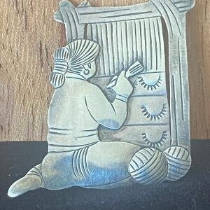 May include: A silver pendant depicting a person weaving on a loom. The pendant is detailed with intricate designs and textures.
