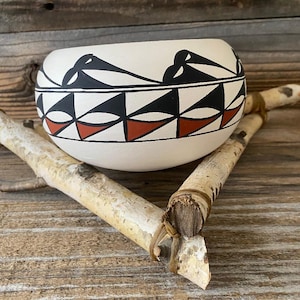 Puede incluir: Un cuenco de cerámica blanca con patrones geométricos negros y rojos. El cuenco tiene forma redondeada y está decorado con un diseño repetitivo de triángulos y figuras estilizadas de pájaros. El cuenco descansa sobre ramas de abedul.