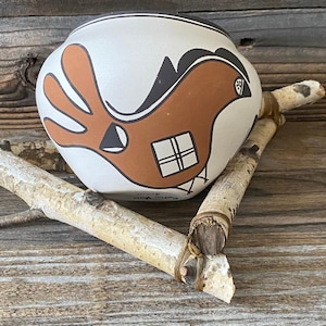 Puede incluir: Una olla de cerámica blanca con un diseño de pájaro marrón y detalles en negro. El pájaro tiene un detalle de ventana cuadrada. La olla descansa sobre una superficie de madera con ramas de abedul.