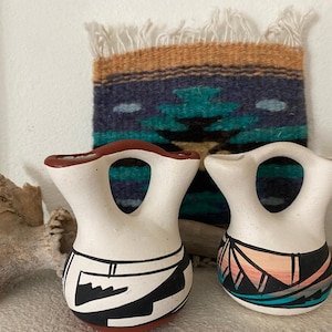May include: Two white ceramic vases with a unique shape and a large opening. One vase has black and white geometric designs, and the other has colorful geometric patterns. A woven tapestry is in the background.