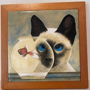 May include: A framed ceramic tile with a painting of a Siamese cat looking at a goldfish in a bowl. The cat has blue eyes and a white and brown coat. The goldfish is orange and swimming in a clear bowl.