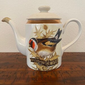 May include: White ceramic teapot with a gold trim and a bird illustration. The bird is brown, yellow, and red with black markings. The bird is perched on a branch with green leaves.