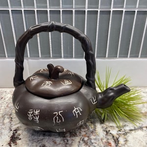 Puede incluir: Una tetera de cerámica marrón con un asa y un pico de estilo bambú. La tetera tiene tapa y está decorada con caracteres chinos blancos. Una ramita de hierba verde está al lado de la tetera.