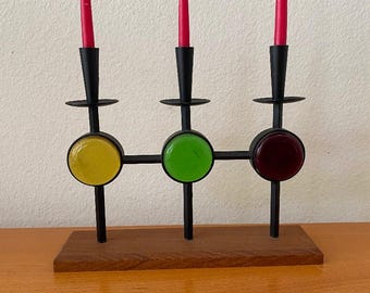 May include: A black metal candle holder with three arms, each with a round colored glass insert. The glass inserts are yellow, green, and red. The candle holder is on a wooden base.