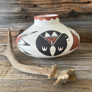 Puede incluir: Un jarrón de cerámica blanco con un borde festoneado y un cuerpo redondeado. El jarrón presenta diseños geométricos negros y rojos y un motivo central de pájaro. Una sección de una cornamenta de ciervo descansa frente al jarrón.