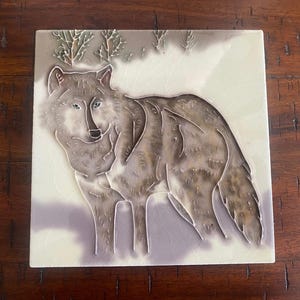 May include: A ceramic tile featuring a raised image of a grey wolf standing in a snowy woodland. The wolf is facing the viewer with its head turned slightly to the side. The tile has a white background with a subtle grey and brown pattern.