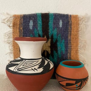 May include: Two handcrafted pottery vases with geometric designs in black and white, and a small orange pot with black and turquoise accents. A woven textile with blue, green, and orange patterns serves as a backdrop.