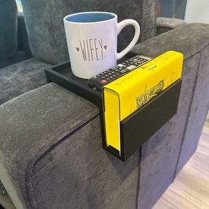 Puede incluir: Bandeja de metal negro para el reposabrazos del sofá con un libro amarillo y una taza blanca con la palabra "WIFEY" en ella.