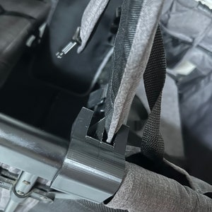 Puede incluir: Primer plano de un asiento de cochecito gris con correas negras y una hebilla de plástico negra. Las correas están sujetas al asiento con un clip de plástico negro.