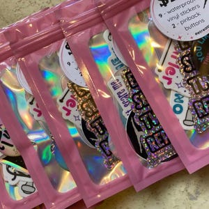 May include: Pink gift bundles with holographic inserts, containing stickers and pinback buttons. The text "Twirl Gift Bundles" is displayed above the bundles. The text "Button styles vary" is at the bottom.