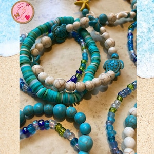 May include: A set of turquoise and white beaded bracelets with turtle charms. The bracelets are layered on a sandy surface.