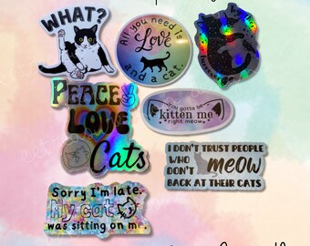 May include: A set of 8 cat-themed stickers with holographic finishes. The stickers feature various cat-related phrases, including "What?", "Peace Love Cats", "All you need is love and a cat", "Sorry I'm late. My cat was sitting on me.", "I don't trust people who don't meow back at their cats", and "You gotta be kitten me right meow."