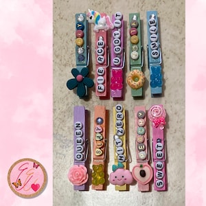 May include: A collection of decorative wooden clothespins in various colors, each adorned with embellishments and words like "FIERCE," "SMILE," and "SWEET." The pins feature charms such as a unicorn, gummy bears, and a donut.