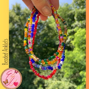 May include: Five colorful beaded anklets in red, orange, yellow, green, blue, and silver. The anklets are layered on top of each other and are held in a hand.
