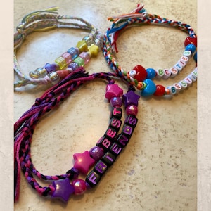 May include: Four handmade friendship bracelets. One bracelet has the words "BEST FRIENDS" spelled out in black letter beads, with pink and purple star-shaped beads. Another bracelet has the word "BESTIES" spelled out in white letter beads. The other two bracelets have colorful beads.