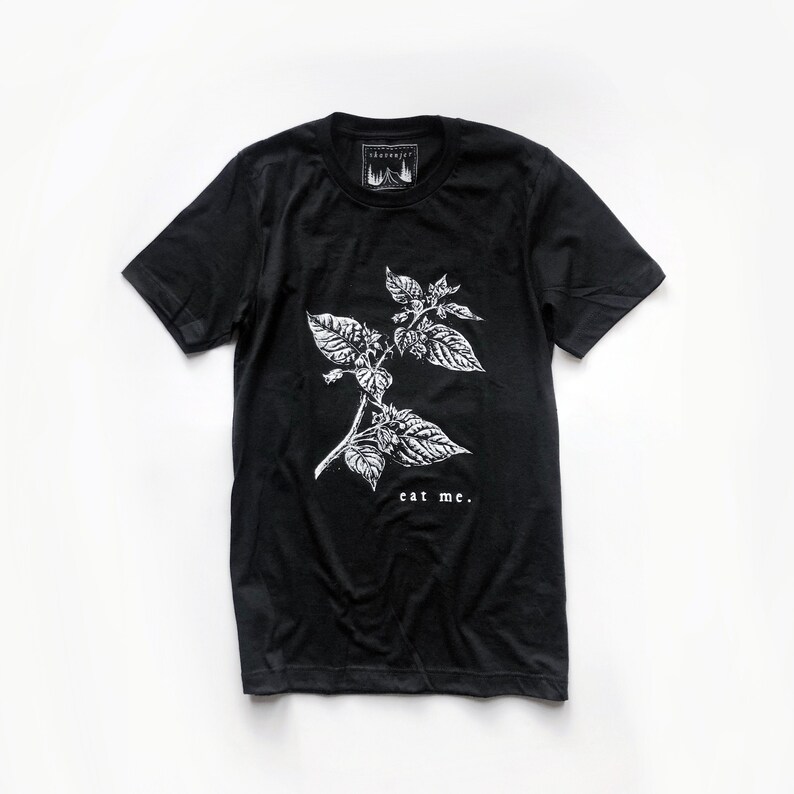 May include: Black t-shirt with a white botanical illustration of a plant and the words "eat me." The shirt has a crew neck and short sleeves. A small tag with a mountain and tree design is at the top.
