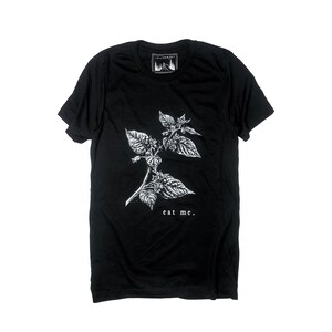 May include: Black t-shirt featuring a white botanical illustration of a plant with the text "eat me." The shirt has a crew neck and short sleeves. A small white label with a mountain and tree design is at the neckline.