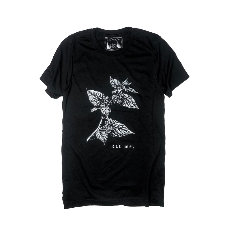 May include: Black t-shirt featuring a white botanical illustration of a plant with the text "eat me." The shirt has a crew neck and short sleeves. A small white label with a mountain and tree design is at the neckline.