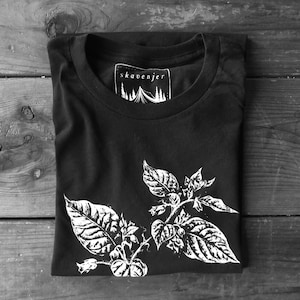 May include: Black t-shirt with a white botanical illustration of leaves and flowers. The shirt has a crew neck and a tag that reads "skavenjer" with a tent and tree graphic. The shirt is folded on a wooden surface.