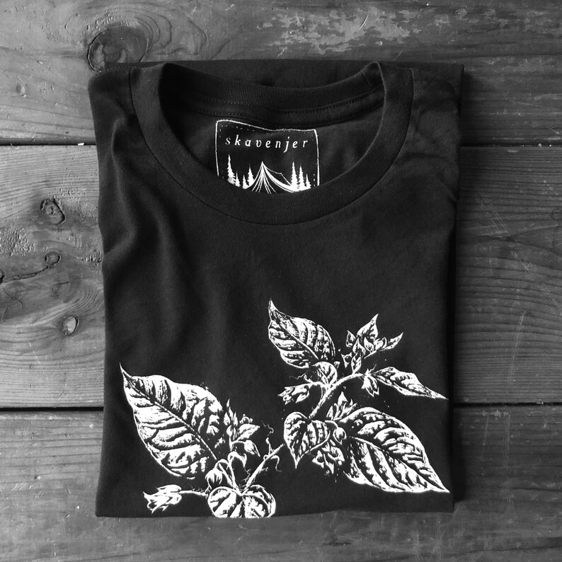 May include: Black t-shirt with a white botanical illustration of leaves and flowers. The shirt has a crew neck and a tag that reads "skavenjer" with a tent and tree graphic. The shirt is folded on a wooden surface.