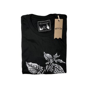 May include: Black t-shirt with a white botanical illustration on the front. The shirt has a crew neck and a tag that reads "skovenjer" with a mountain and tent graphic. A brown tag with "SKVJR" hangs from the shirt.