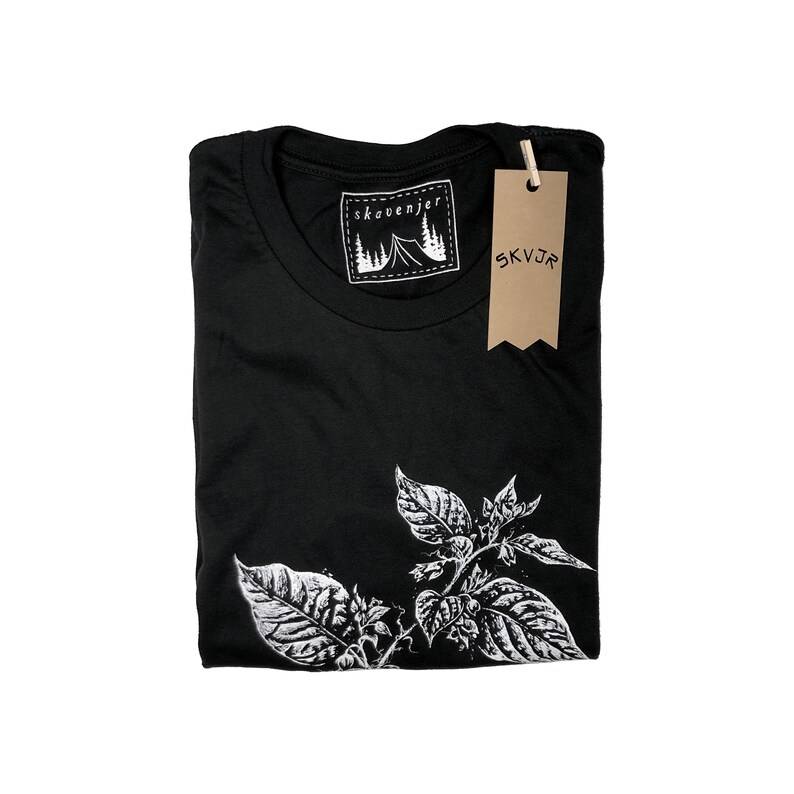 May include: Black t-shirt with a white botanical illustration on the front. The shirt has a crew neck and a tag that reads "skovenjer" with a mountain and tent graphic. A brown tag with "SKVJR" hangs from the shirt.