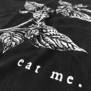 May include: Black t-shirt featuring a detailed white illustration of basil leaves and stems. The phrase "eat me" is embroidered in white at the bottom. The design is centered and takes up most of the shirt's surface.