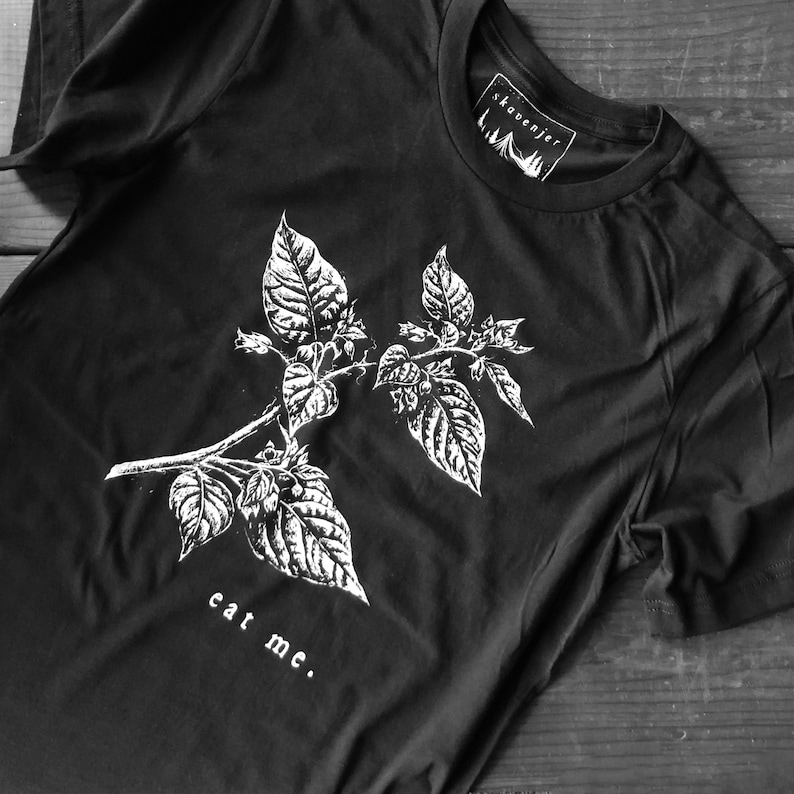 May include: Black t-shirt featuring a white botanical illustration of a plant with large leaves and the words "eat me." The shirt has a crew neck and a tag that says "skavenjer."