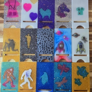 May include: A collection of fifteen rectangular resin phone stands featuring various designs including medical scrubs, hearts, animals, space themes, and silhouettes. Each stand has a slot for a device and a cutout at the bottom for charging cables.