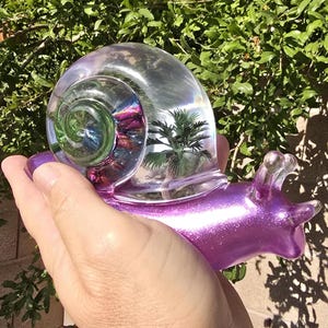 May include: A glass snail figurine featuring a glittery purple body and a clear shell. The shell contains a swirling green and magenta center with a small green palm tree encased inside. The snail has two clear antennae and a smooth, shiny finish.