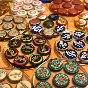 May include: A collection of circular coasters made of clear resin, each embedded with various metal bottle caps from different beverage brands. The caps feature diverse logos, colours, and text, including Corona Extra, Heineken, Kopparberg, and Amstel, arranged in clusters.