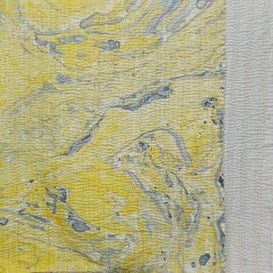 May include: Close-up of a textile with a marbled pattern in shades of yellow and gray. The fabric has a textured surface with a wavy, rippled appearance. A section of white, textured fabric is visible on the right side.