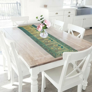 Op de afbeelding: Een turquoise en gouden tafelkleed met een bloemenpatroon ligt op een witte houten tafel met witte stoelen. Een vaas met roze bloemen staat op het tafelkleed.