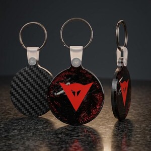 Könnte beinhalten: Drei kreisförmige Schlüsselanhänger mit silbernen Metallringen und weißen Kunststoffverbindern. Einer zeigt ein schwarzes Carbonfasermuster, während die anderen beiden ein schwarz-rotes Glitzer-Finish mit einem roten dreieckigen Logo in der Mitte aufweisen.