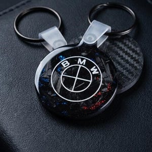 Könnte beinhalten: Ein runder Schlüsselanhänger mit dem weißen BMW-Logo auf einem schwarzen, strukturierten Hintergrund in Carbon-Optik mit eingearbeiteten blauen und roten Sprenkeln. Das Emblem ist in eine glänzende, klare Harzschicht eingebettet.