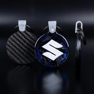 Könnte beinhalten: Drei kreisförmige Schlüsselanhänger mit Metallringen und durchsichtigen Kunststofflaschen. Einer hat ein schwarzes Carbonfaser-Muster, ein anderer zeigt ein weißes Suzuki-Logo auf blauem Glitzergrund und der dritte zeigt das Profil der Scheibe.