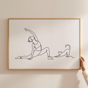 Puede incluir: Una impresión enmarcada con un dibujo lineal minimalista de una persona en una postura de yoga con un gato estirándose. La obra de arte es en negro sobre un fondo crema, dentro de un marco de madera marrón claro.