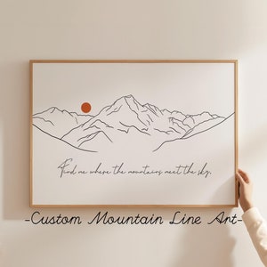 以下が含まれることがあります： 背景に赤い太陽が描かれた、ミニマルな線画の山のプリント。山の下には「Find me where the mountains meet the sky.」というテキストが書かれています。プリントは明るい木製のフレームで額装されています。