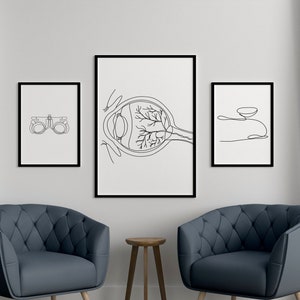 May include: Three black framed illustrations of the human eye, an eye chart, and a contact lens. The illustrations are hanging on a white wall between two blue curtains. Two blue armchairs with wooden legs are in front of the illustrations.
