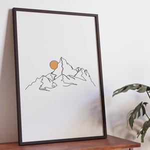 Puede incluir: Impresión artística enmarcada con un dibujo lineal minimalista de una cordillera y un sol dorado. La obra de arte se exhibe sobre una mesa de madera. Un jarrón blanco y una planta verde están en el fondo. La impresión añade un toque de naturaleza.