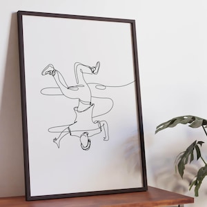 May include: Black framed art print featuring a continuous line drawing of a person doing a handstand. The artwork is minimalist, with the figure rendered in a single, unbroken black line against a white background. The print is displayed on a wooden table.