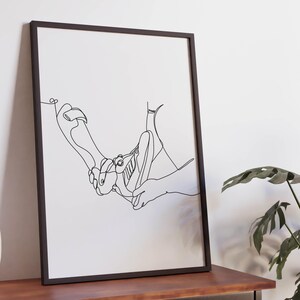 Puede incluir: Lámina artística enmarcada en negro con un dibujo de línea continua de dos manos sosteniendo un zapato. La obra de arte se exhibe sobre una mesa de madera oscura contra una pared blanca, con un jarrón decorativo y una planta en maceta en primer plano.