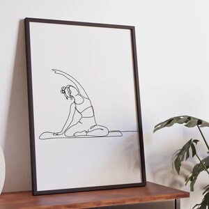 May include: A framed black and white line art print of a person in a yoga pose. The artwork shows a seated figure with one arm raised overhead. The print is displayed on a wooden table with a decorative vase and a potted plant.
