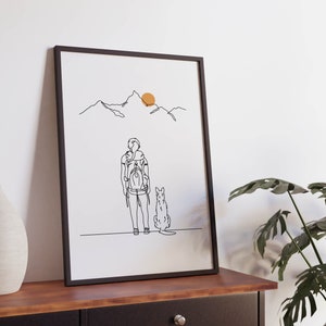 May include: A black framed print of a line drawing of a person with a backpack and a dog standing in front of a mountain range with a yellow sun in the sky.