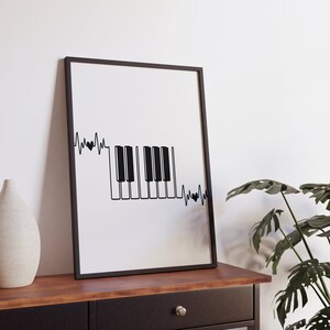 Peut inclure: Impression en noir et blanc d'un clavier de piano avec une ligne de battement de cœur au-dessus et en dessous. L'impression est encadrée dans un cadre noir et est accrochée à un mur blanc au-dessus d'une commode en bois.